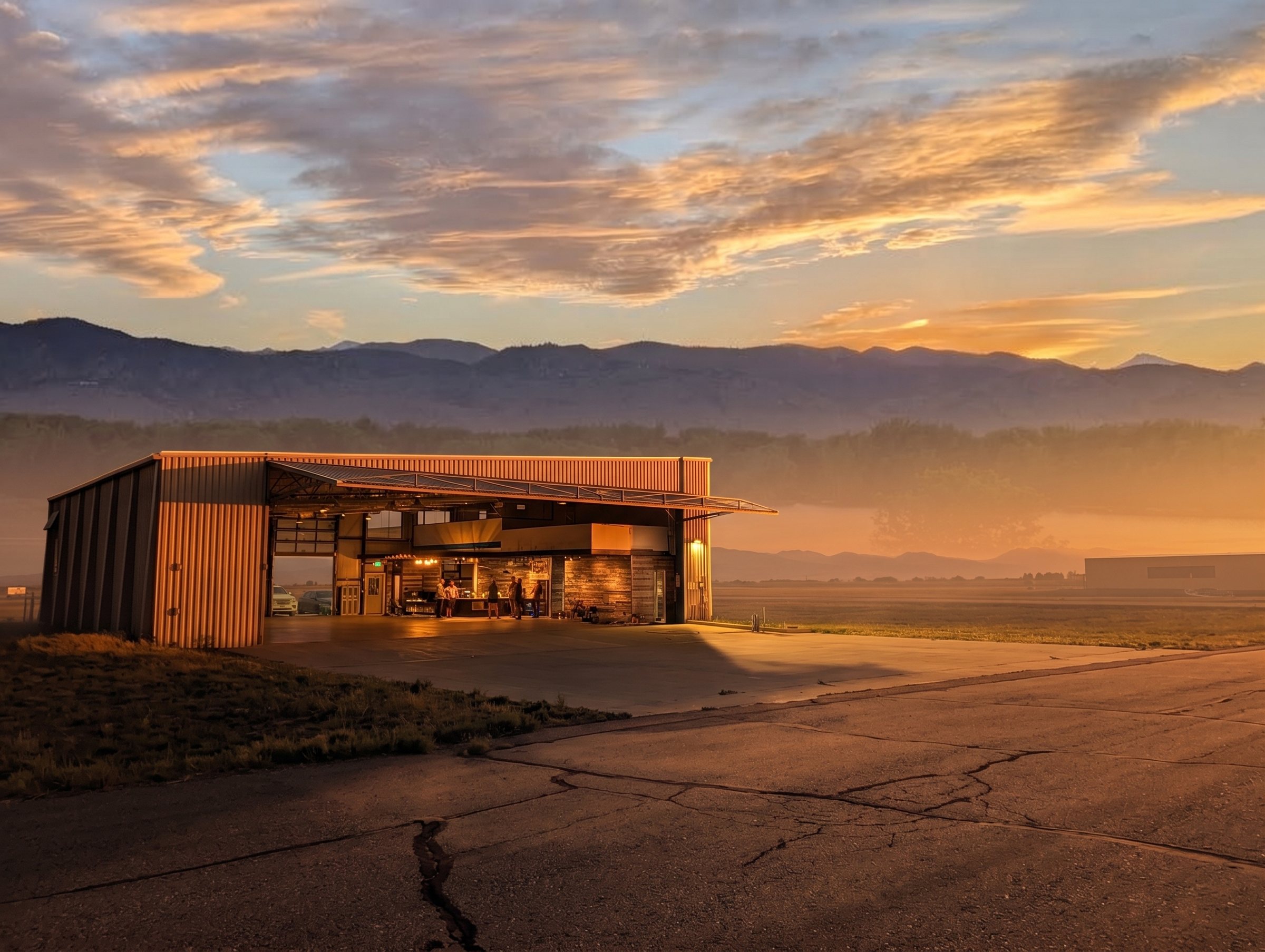 The building at golden hour, doors open and the bar lit, with the Front Range behind it.